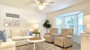 A spacious living room with a white sofa, two armchairs, a coffee table, and a ceiling fan. There are framed pictures on the wall and a large window providing natural light.
