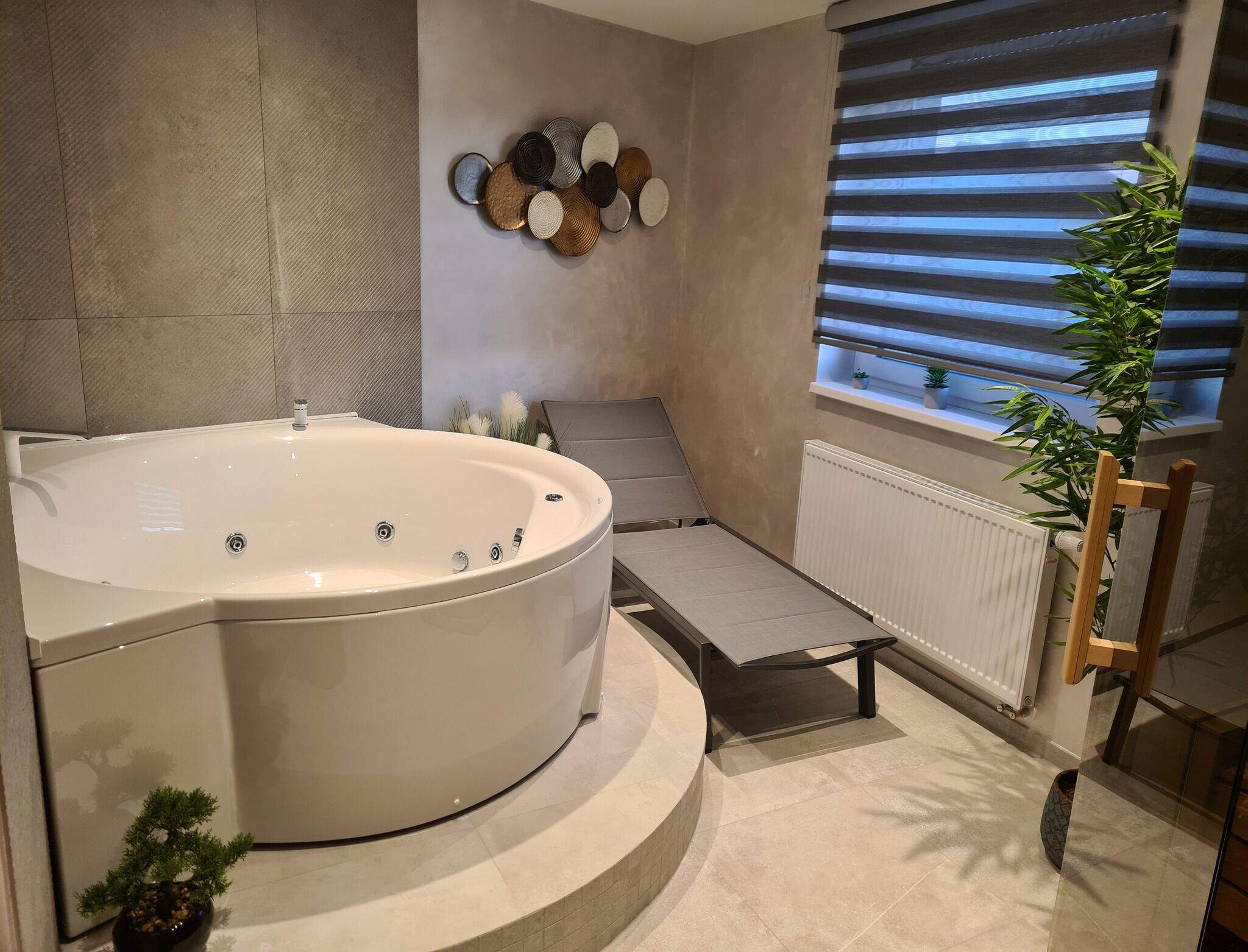 A modern bathroom with a large bathtub, a small seating area, a radiator, and a window with blinds.