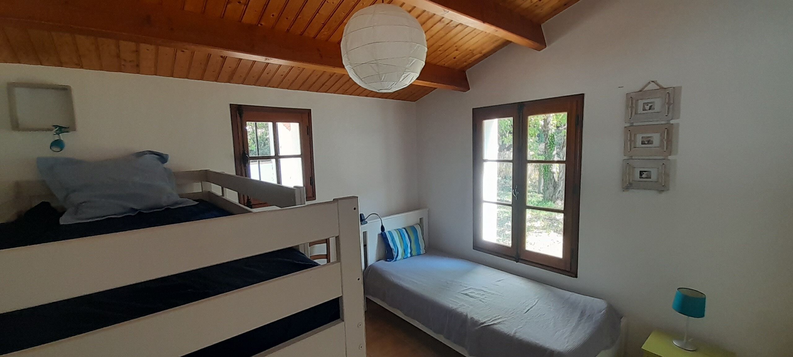 A room with a bunk bed, a single bed, a window with a view of greenery, a hanging light fixture, and a wall-mounted picture frame.