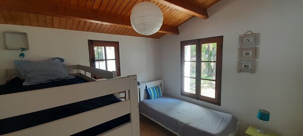 Habitació amb un llit superposat, un llit individual, una finestra que dona a un espai verd, una làmpada penjant i un quadre penjat a la paret.