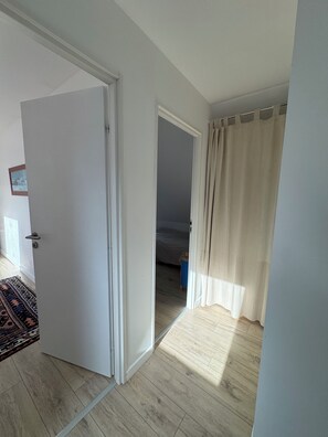 A room with a wooden floor, a bed with a striped bedspread, a blue trash bin, and a beige curtain. - MEZOU BRAS (LANDUNVEZ)