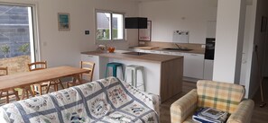 Une cuisine compacte avec un plan de travail en bois, un évier et un petit réfrigérateur. On y trouve une table à manger avec des chaises, ainsi qu’un canapé dans l’espace salon.