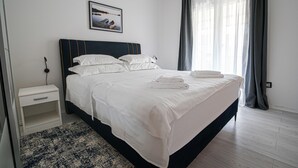 Een netjes opgemaakt bed met witte lakens en opgevouwen handdoeken, een nachtkastje met een lamp, een ingelijste foto aan de muur en een raam met gordijnen.