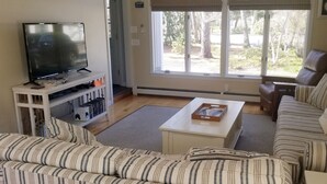 Un salon avec un téléviseur posé sur un meuble blanc, un canapé, une table basse et une fenêtre donnant sur des arbres.