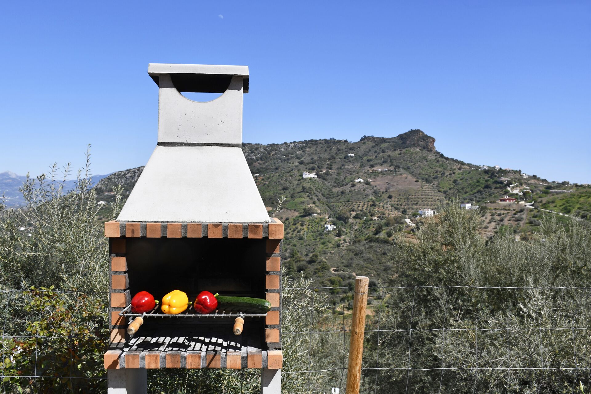 Una parrilla de ladrillo al aire libre con chimenea, rodeada por una valla de alambre y vegetación, ubicada en una ladera con casas dispersas.