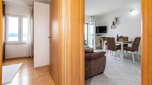 En moderne stue med sofa, spisebord med stole, fjernsyn og havudsigt gennem et vindue.