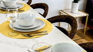 Una mesa de comedor dispuesta con platos, tazones y cubiertos blancos, rodeada de sillas.