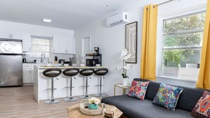 Una cocina moderna con armarios blancos, un refrigerador de acero inoxidable y una zona de bar con cuatro taburetes. Hay un sofá gris con cojines coloridos y una pequeña mesita de madera con un florero y una bandeja.
