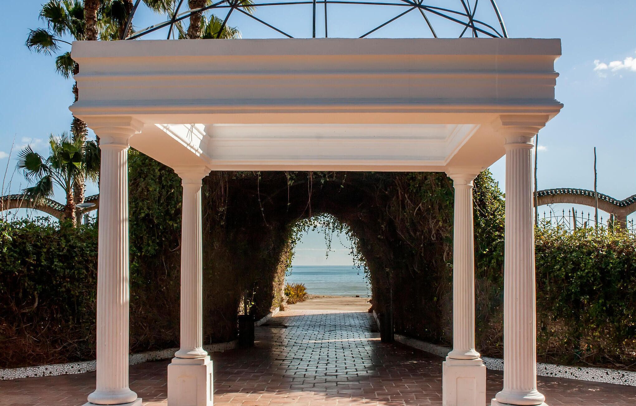 A white pergola with columns, leading to a beach path.