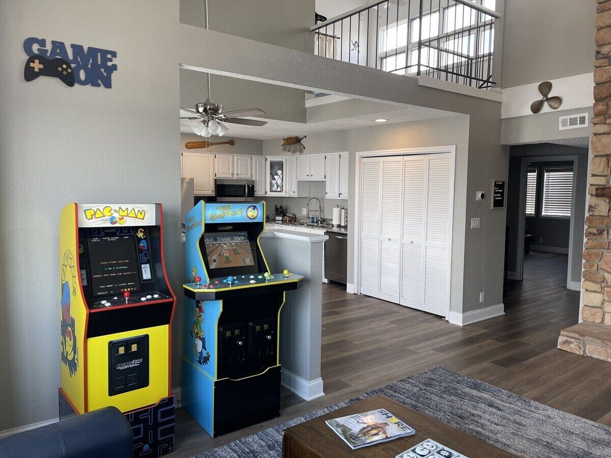 Una sala de estar con dos máquinas de videojuegos, una cocina al fondo y una escalera que lleva a una zona tipo loft.