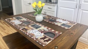 Una mesa de madera para comer con un mantel decorativo que presenta personajes de caricaturas y texto. Un jarrón blanco con tulipanes amarillos está colocado sobre la mesa.