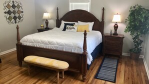 Un dormitorio con una cama de madera, dos mesitas de noche con lámparas, un banco, un adorno en la pared y una planta en maceta.