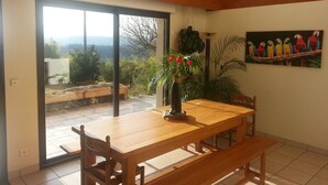 A dining area with a wooden table and chairs, a view through a glass door to an outdoor patio, and a bookshelf with books.