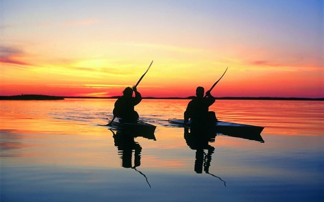 Dos personas remando en canoas sobre aguas tranquilas al atardecer.