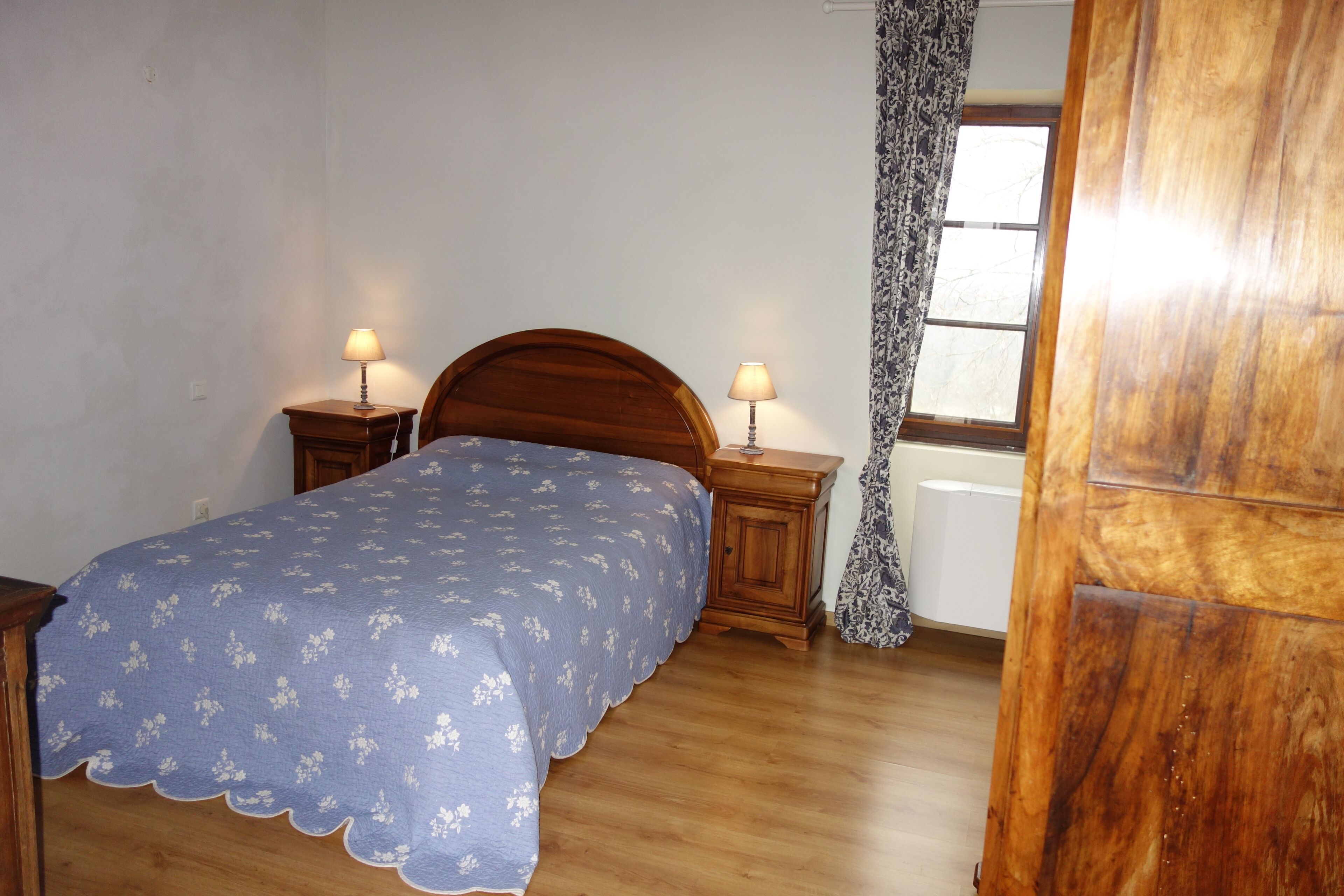 A bedroom with a wooden bed, two bedside tables with lamps, a window with a curtain, and a radiator.