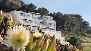 Un immeuble moderne à plusieurs étages, doté de balcons, entouré d’une végétation luxuriante et de collines.