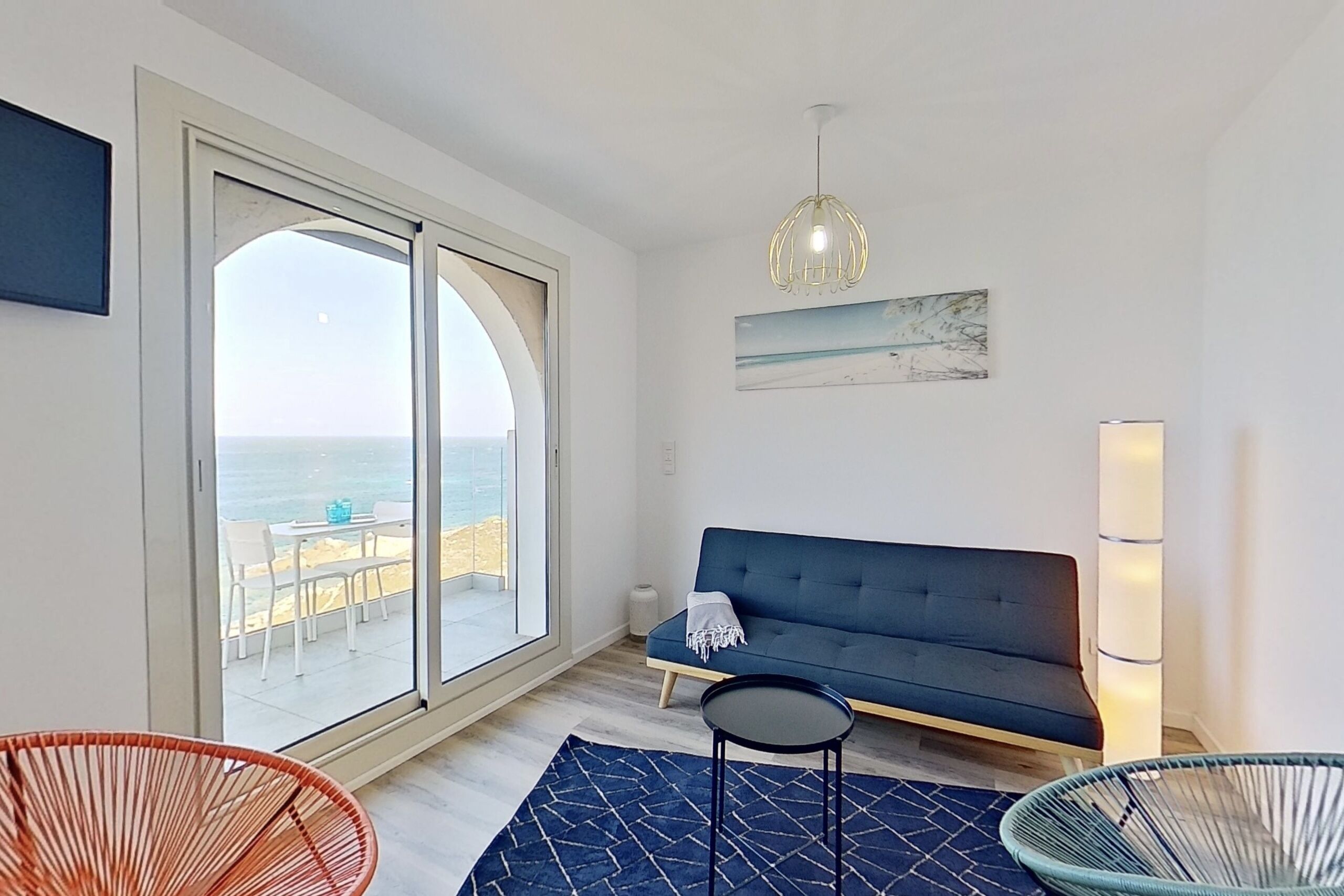 A modern living room with a sofa, a small table, and a chair, a large window with a view of the beach, and a hanging light fixture.
