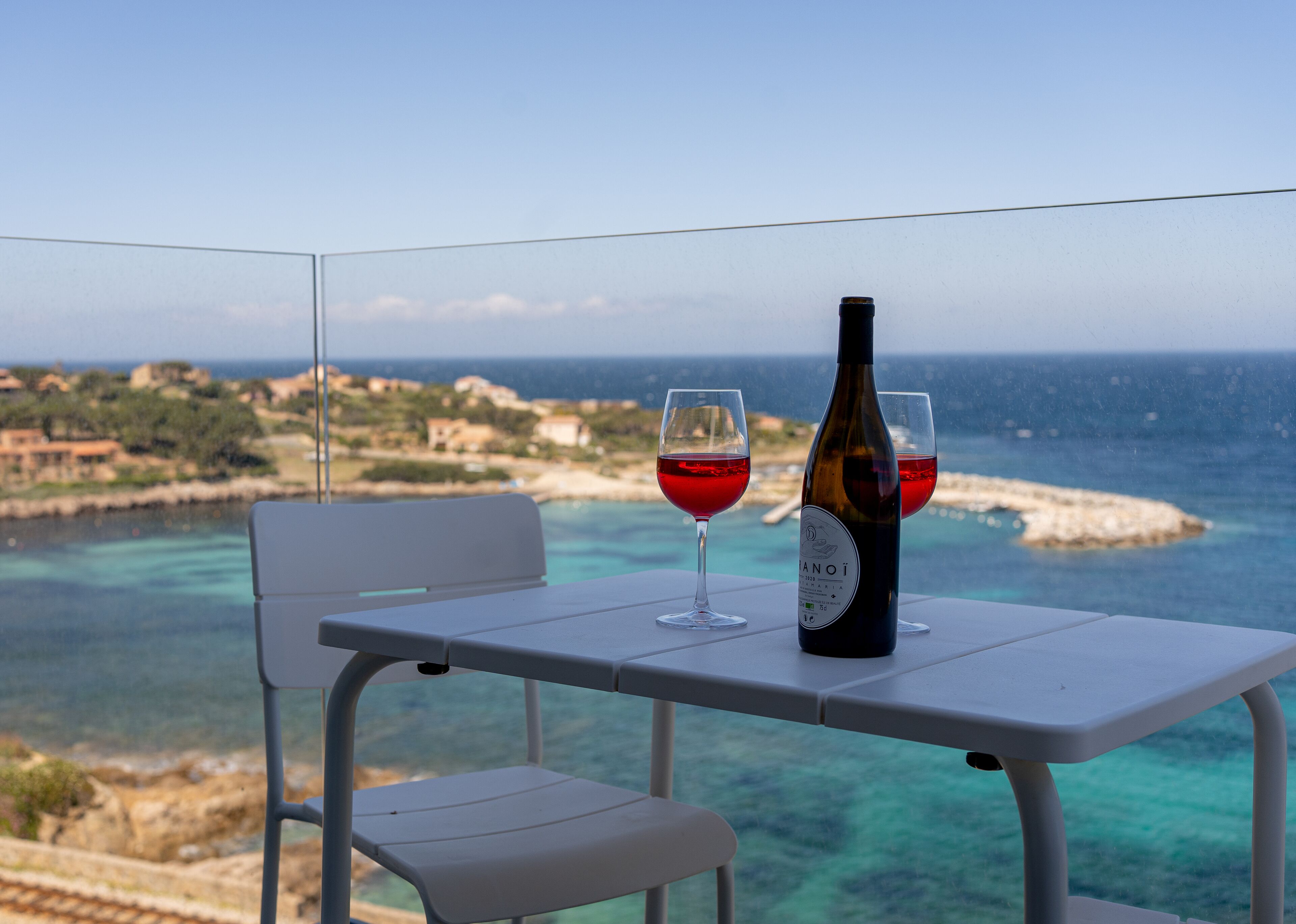 A table with a bottle and two glasses of red wine, overlooking a coastal landscape.