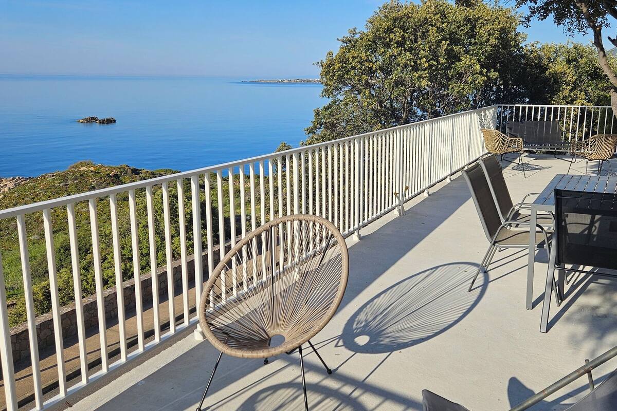 Un balcon avec vue sur la mer, des fauteuils en osier, une table et un arbre.