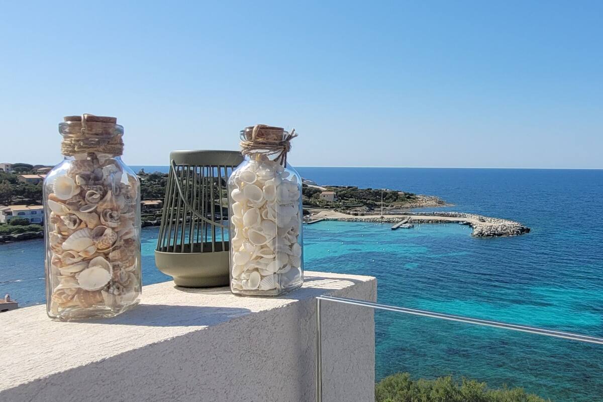 Deux bouteilles en verre remplies de coquillages sont posées sur un balcon donnant sur une zone côtière, avec une eau d’un bleu limpide et une côte rocheuse.