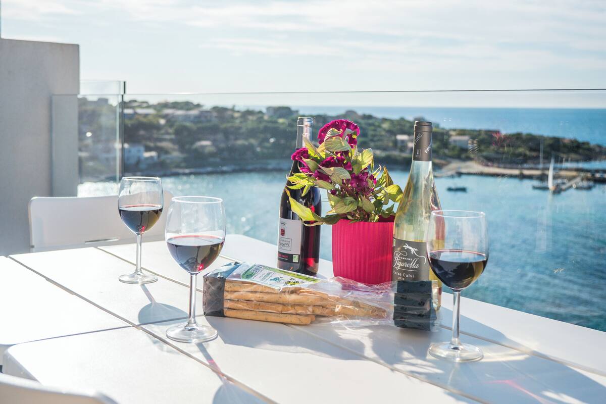 Une table avec des verres à vin, une bouteille de vin, un bouquet de fleurs et un livre, le tout placé sur un balcon donnant sur une étendue d’eau.