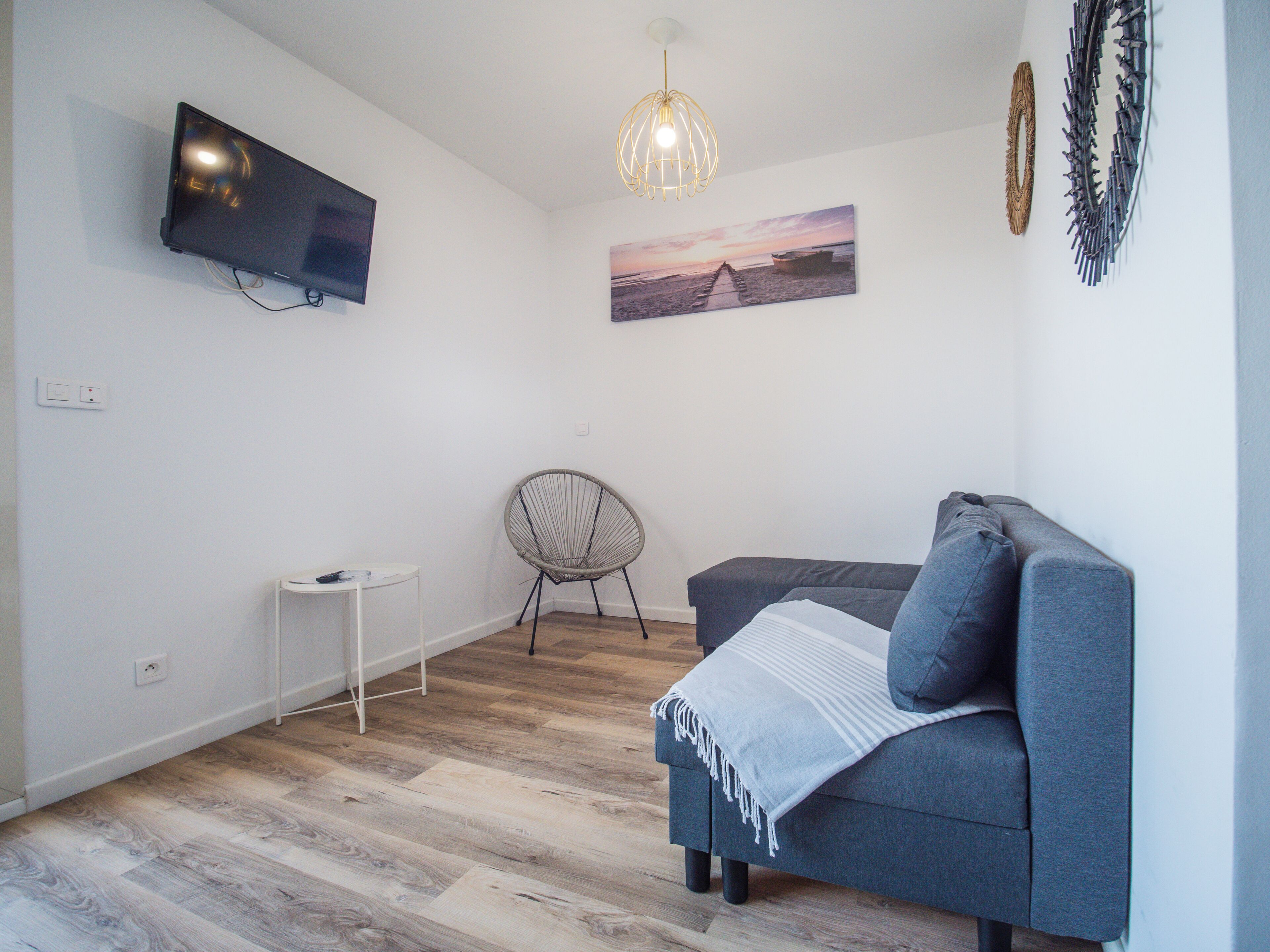 A modern living room with a grey sofa, a small white side table, a wicker chair, a mounted TV, and a wall-mounted picture.