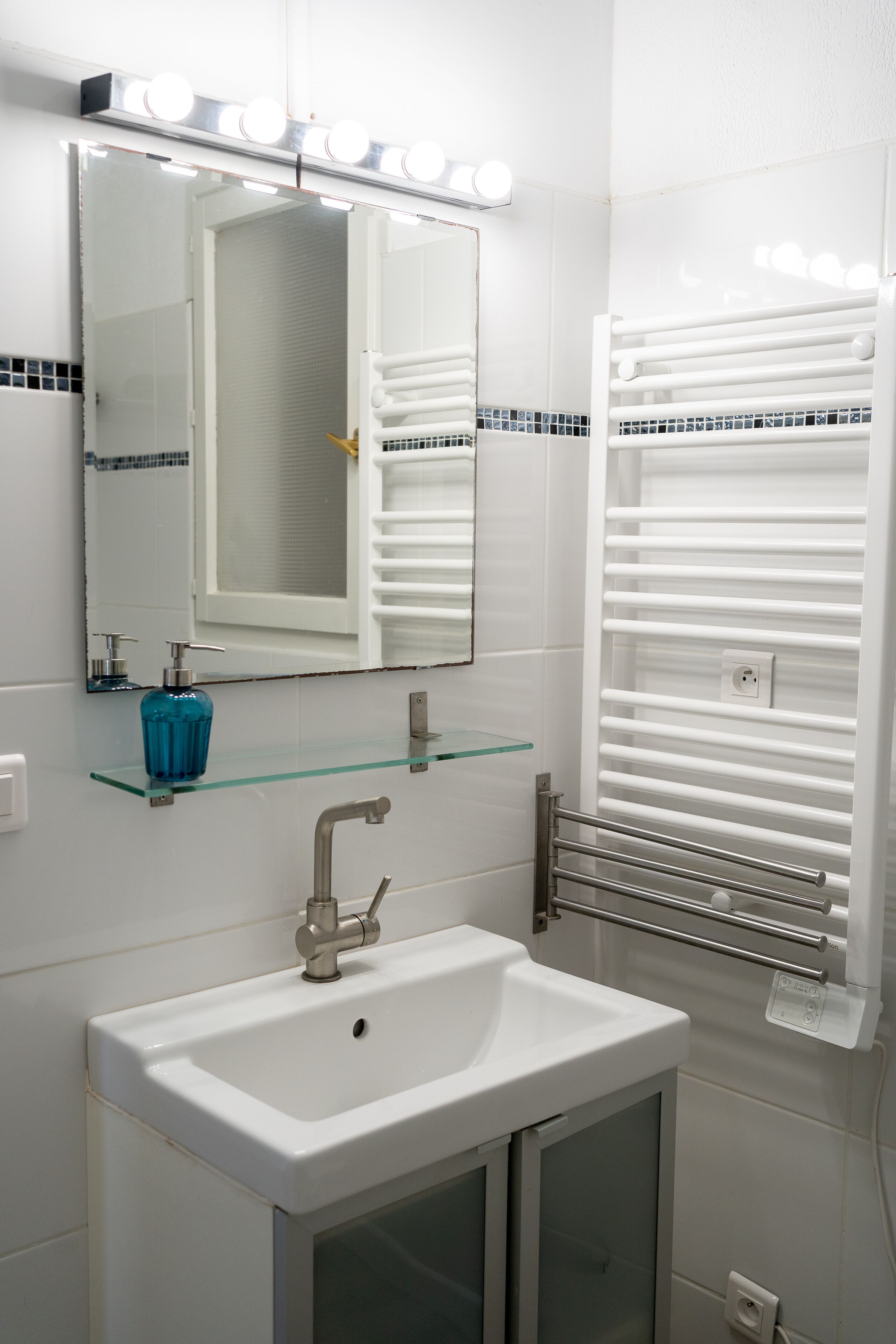 Un baño con un lavabo blanco, un estante de cristal con una botella azul, un espejo con iluminación y un perchero para toallas.