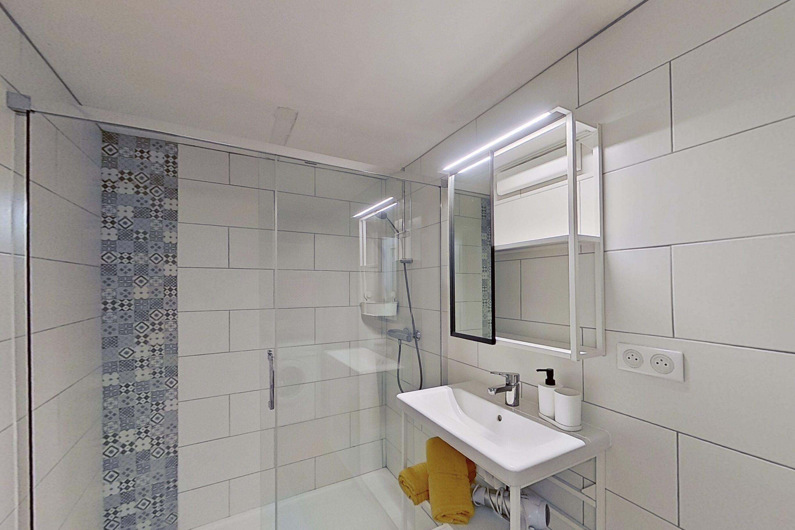 A modern bathroom with a glass shower enclosure, a white sink with a mirror, and patterned tiles on the walls.