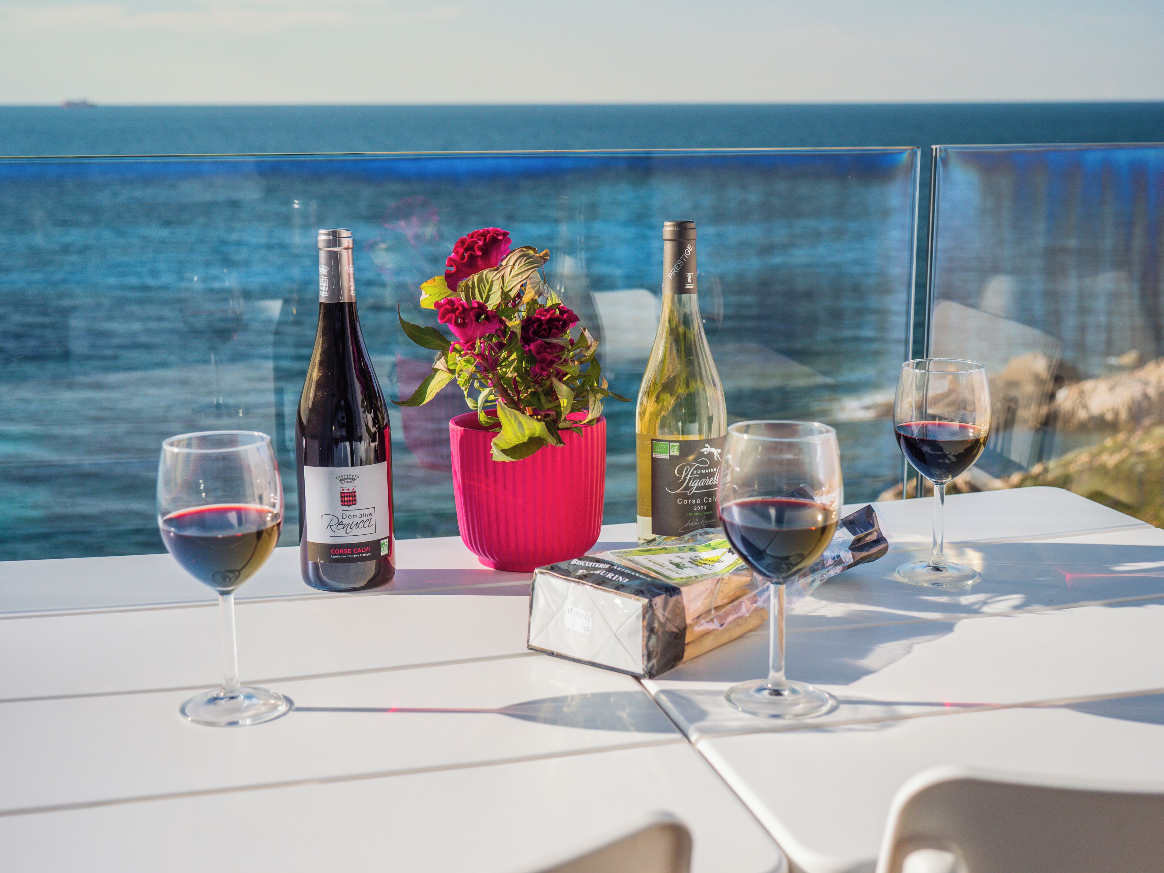 A table with wine bottles and glasses, a pink vase with flowers, and a view of the sea.