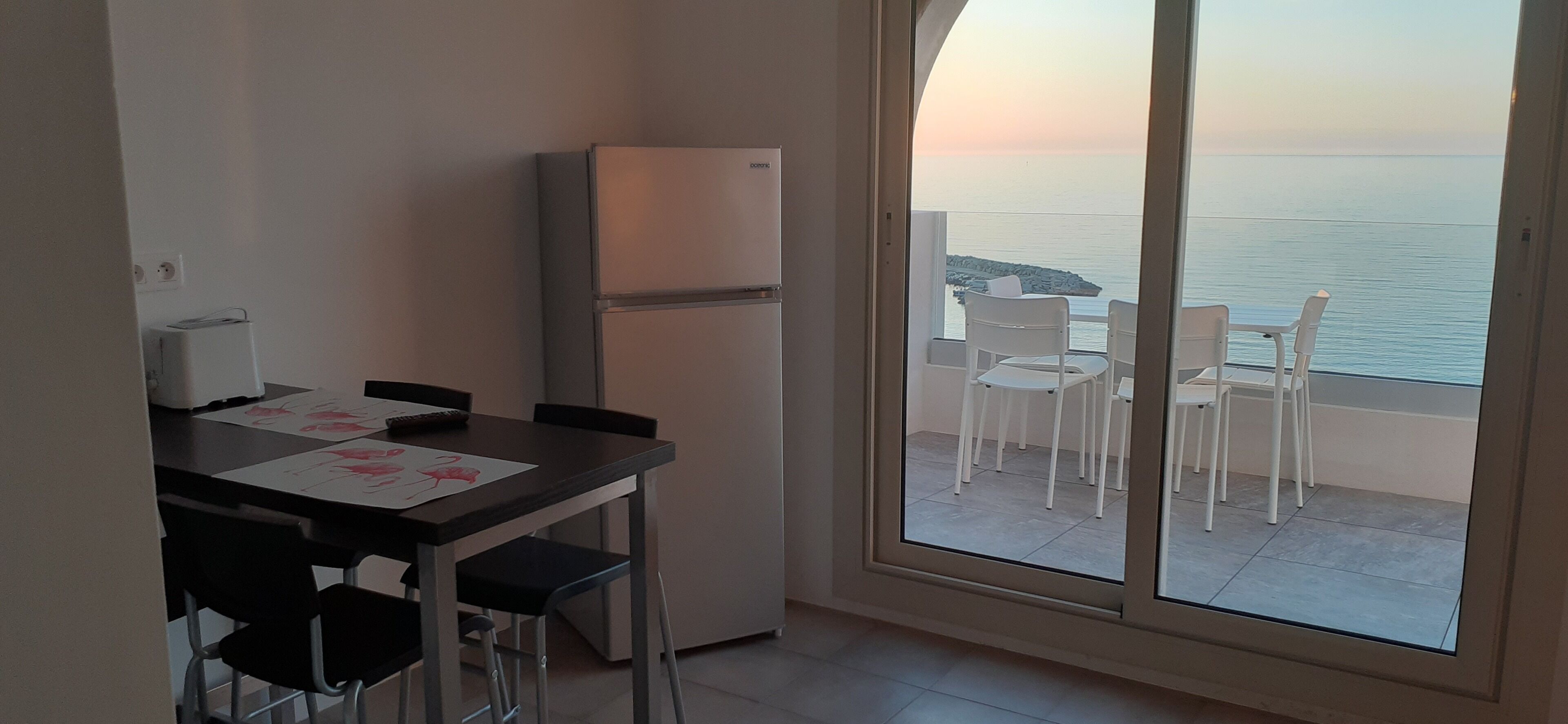 A compact kitchen with a refrigerator, a dining table with chairs, and a large sliding glass door leading to a balcony with a view of the sea.