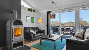 A modern living room with a wood-burning stove, a black leather sofa, a coffee table, and a view of the outdoors through large windows. - Gorgeous home in Blokhus with sauna (Blokhus)