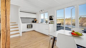 Uma cozinha moderna com armários brancos, piso de madeira e grandes janelas com vista para um rio e vegetação.