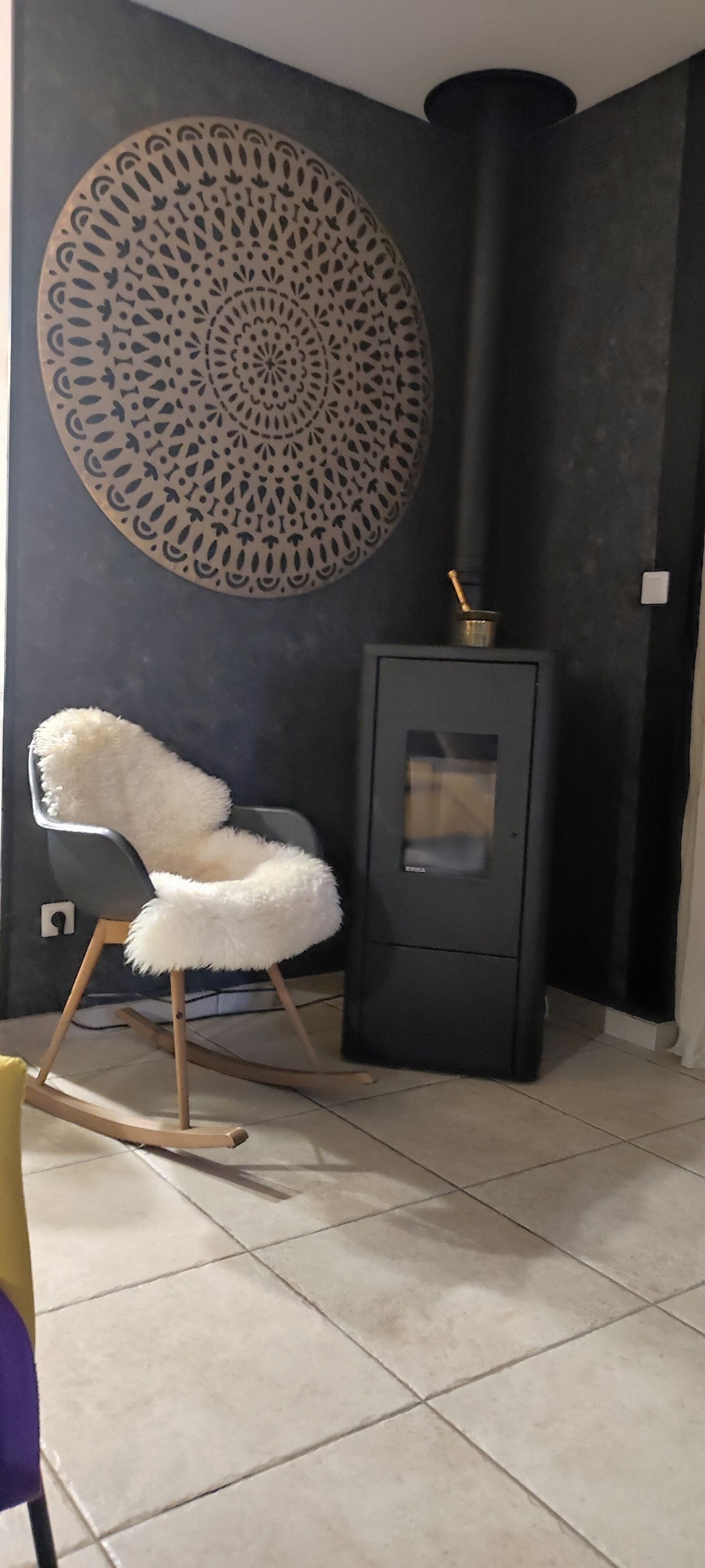 Una habitación con un adorno circular en la pared, un sillón blanco y mullido, un mueble negro con estufa y suelo de baldosas.