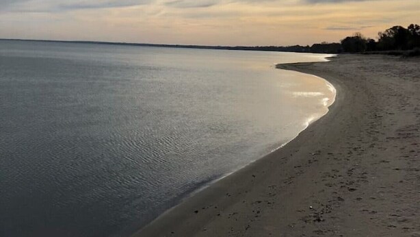 A beach with a calm sea, a sandy shore, and a cloudy sky with the sun visible. - Sandy Neighborhood Private Beach Getaway in Huron (Huron)
