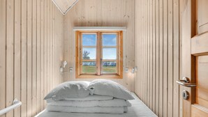 A narrow room with wooden walls, a bed with white bedding, a window with a view of the outdoors, and a door on the right.