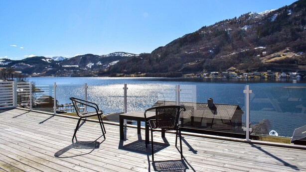 Una terraza de madera con dos sillas y una mesa, con vista a un lago y montañas al fondo.