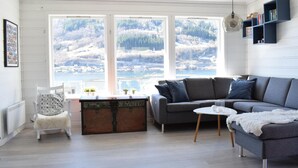 Un salón acogedor con un sofá gris, una mesita redonda y vistas a un lago y montañas a través de grandes ventanales.