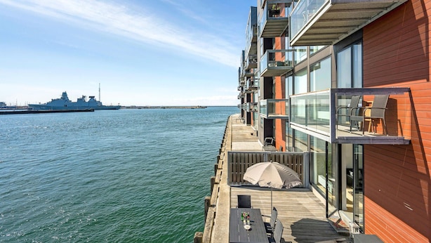 A modern apartment with a balcony overlooking a body of water, a ship in the background, and a view of a distant city skyline. - Amazing apartment in Korsør (Korsør)