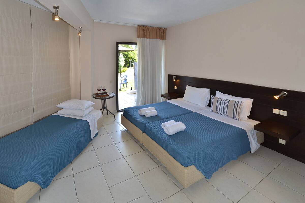 A hotel room with two single beds, a small table with two glasses of red wine, and a view of the outdoor area through a glass door.