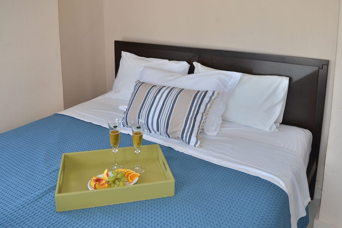 A neatly made bed with white linens and striped pillows, a blue textured bedspread, and a green tray with fruit and drinks.