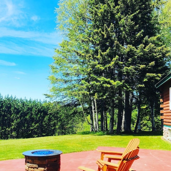 A wooden deck with a stone fire pit, a wooden Adirondack chair, and a view of a forest with tall trees.