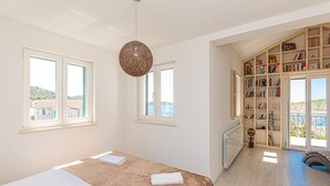 Ein Schlafzimmer mit einem Bett, zwei Fenstern, einer Pendelleuchte, einem Bücherregal und Meerblick.