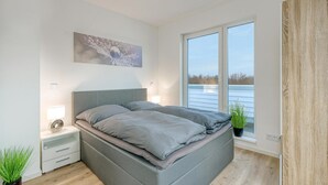 Ein modernes Schlafzimmer mit einem großen Bett, einem Nachttisch, einer Nachttischlampe, einem Fenster mit Blick auf ein Gewässer und einer Zimmerpflanze.