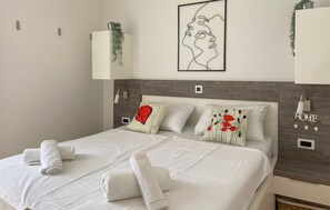 Ein ordentlich bezogenes Bett mit weißen Leinen und gerollten Handtüchern, ein Kopfteil mit Holzstruktur und ein an der Wand hängendes Kunstwerk, das ein Frauenprofil zeigt.