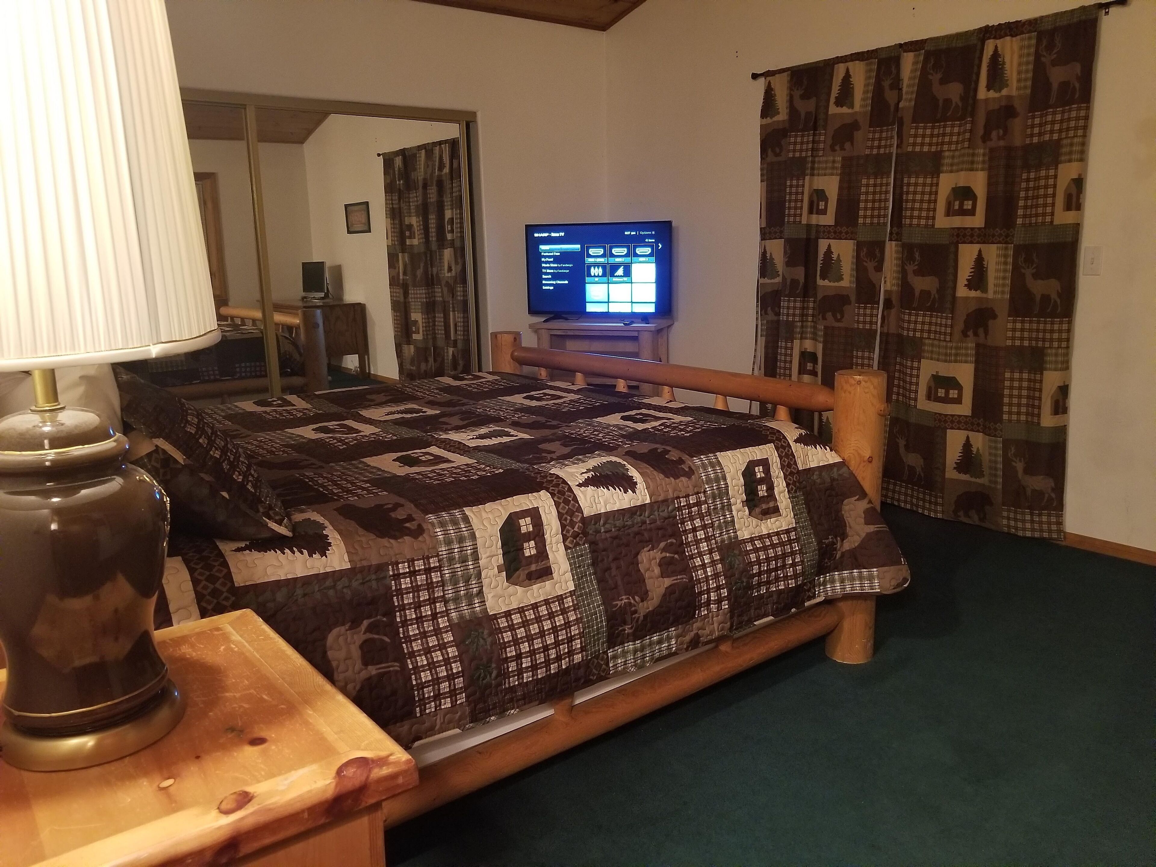 Ein Schlafzimmer mit einem hölzernen Bett, einem Fernseher auf einem Schrank, einer Lampe und gemusterten Vorhängen.