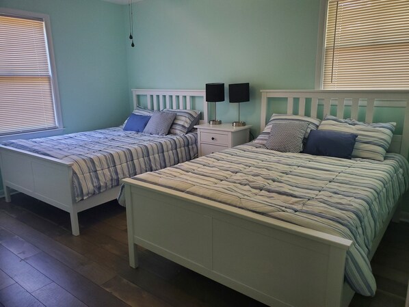 A bedroom with two single beds, a nightstand with two lamps, and a window with blinds. - Sandy Paws - A Dog-Friendly Haven with Boat Parking on Oak Island, NC (Oak Island)