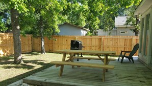 Un jardin avec une terrasse en bois, une table de pique-nique, un barbecue et des chaises Adirondack.