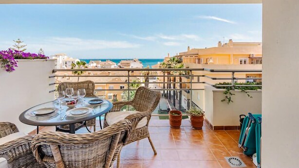 Un balcón con mesa lista para comer, mobiliario de mimbre, plantas en macetas y vista al mar y edificios.