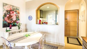 Un comedor con una mesa redonda, sillas y un jarrón de flores. Hay un reloj en la pared y una puerta que da a la cocina.