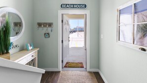 Een gang met een 'Beach House' bord, een spiegel, een klein bureau en een deur naar buiten.